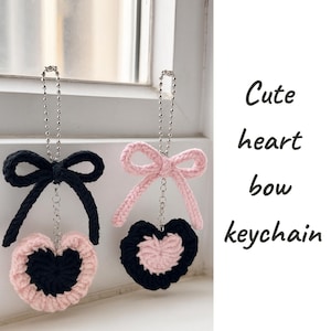 Può includere: Due portachiavi a forma di cuore all'uncinetto. Un portachiavi ha un fiocco nero e un cuore rosa e nero. L'altro ha un fiocco rosa e un cuore nero e rosa. Il testo a destra dice "Cute heart bow keychain."