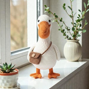 May include: A handmade, crocheted goose doll with a white body, orange beak and feet, and a small brown purse. The goose has black eyes and is standing on a windowsill. A small succulent plant in a pot and a vase with greenery are also visible.