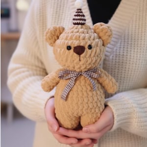 Puede incluir: Un oso de peluche tejido en color beige con nariz y ojos marrones, que lleva un gorro de fiesta con un pompón blanco. El oso tiene un lazo a cuadros marrón y blanco. El oso está siendo sostenido en las manos de alguien.