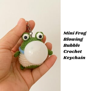 May include: A green crochet frog keychain with large black eyes and a white bubble. The frog has a blue collar and a cream-colored belly. The text "Mini Frog Blowing Bubble Crochet Keychain" is on the right.