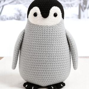May include: A gray crocheted penguin stuffed animal with black eyes, beak, and feet. The penguin is sitting on a white surface.