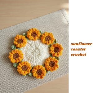 Könnte beinhalten: Ein handgefertigter Häkeluntersetzer in Form einer Sonnenblume. Der Untersetzer hat eine weiße Mitte, umgeben von gelben Blütenblättern mit braunen Zentren und grünen Blättern. Der Text "sunflower coaster crochet" ist sichtbar.