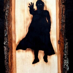 May include: A wooden plaque with a dark silhouette of a figure reaching out. The figure is wearing a long coat and has glowing white eyes. The wood has a natural bark edge and a light brown center.