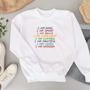 May include: White sweatshirt with a rainbow-colored message that reads "I am kind, I am smart, I am brave, I am confident, I am capable, I am grateful, I am loved, I am enough."