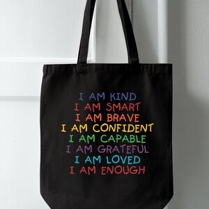 May include: Black tote bag with the text "I am kind, I am smart, I am brave, I am confident, I am capable, I am grateful, I am loved, I am enough" in rainbow colors.