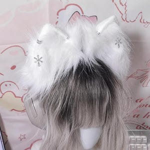 May include: A pair of white faux fur cat ears attached to a headband, featuring silver snowflake charms and silver rings on each ear. The ears are positioned on top of a mannequin head with long, wavy, light-colored hair and bangs.
