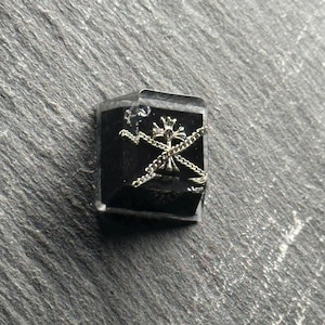 May include: A square black mechanical keyboard keycap featuring a centered silver-toned cross emblem. Two thin silver-toned metal chains are arranged in an overlapping X pattern across the front face of the keycap, partially obscuring the cross.