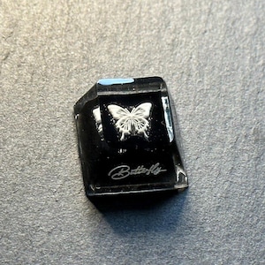 May include: A black mechanical keyboard keycap featuring a white butterfly graphic centered on the top surface and the word Butterfly written in a white script font along the lower edge of the keycap face, with a glossy finish throughout.