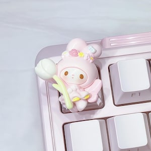 May include: A light pink mechanical keyboard with white keycaps. A decorative pink figurine of a cartoon character holding a white flower is placed on the keyboard. The keyboard has a light pink frame.