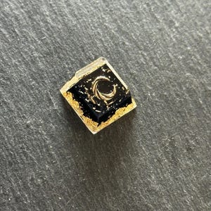May include: A square mechanical keyboard keycap featuring a deep black resin base with embedded gold leaf flakes and a prominent metallic gold crescent moon symbol centered on the top surface, encased in a clear, glossy resin finish.