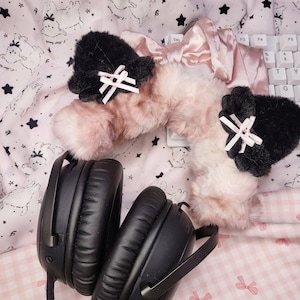 May include: A pair of black over-ear headphones featuring a plush, fuzzy headband cover in a light pink and white gradient. The headband is adorned with two black faux fur cat ears, each decorated with a small, pale pink ribbon bow.