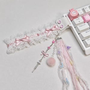 May include: A white lace wrist cuff featuring pink satin bows, attached to a white computer keyboard. A silver chain charm hangs from the cuff, adorned with a small metal syringe, a pink heart bead, a pink pom-pom, and pastel fabric ribbons.