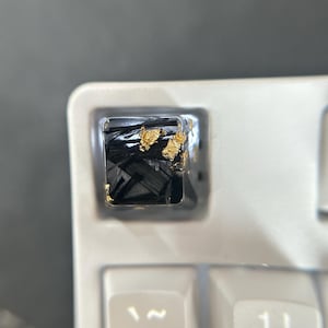 May include: Close-up of a keyboard key with a unique design. The keycap features a black, textured interior with gold leaf accents, encased in a clear resin. The surrounding keys are a light, neutral color.