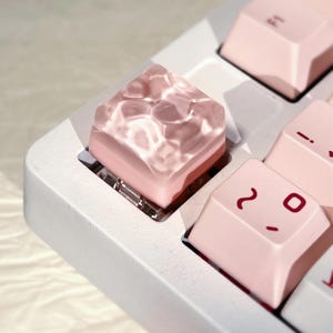 Könnte beinhalten: Nahaufnahme einer individuellen Tastatur mit einer einzigartigen Taste. Die Taste ist ein durchscheinender, strukturierter Würfel mit rosa Basis, der Wasser ähnelt. Andere Tasten sind hellrosa mit dunkelroten Symbolen, auf einer weißen Tastatur.