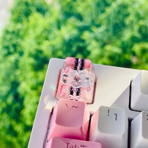 Puede incluir: Una tecla de teclado personalizada con un diseño rosa y blanco, con un lazo decorativo y una ilustración detallada. La tecla está colocada en un teclado blanco con teclas rosas y blancas, sobre un fondo verde borroso.