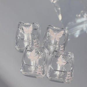 May include: Four clear, square-shaped mechanical keyboard keycaps featuring a light pink butterfly graphic, a small silver star accent, and a pink lace-up corset-style detail printed on the front surface of each individual keycap.