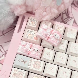 May include: A light pink mechanical keyboard featuring custom keycaps with illustrations of cakes, bows, and a character with long ears. The keys include text such as Shift, Win, and symbols like #3, all decorated with delicate floral patterns and soft pink accents.