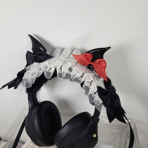 Peut inclure: Casque audio noir orné d'un serre-tête décoratif avec des oreilles noires en forme de chauve-souris, une bordure en dentelle blanche et des nœuds rouges. L'accessoire est fixé sur le dessus du casque.