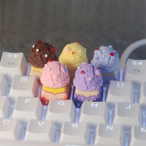 Puede incluir: Primer plano de un teclado blanco con teclas personalizadas en forma de conos de helado coloridos. Las teclas son marrones, amarillas, azules, rosas y moradas, con detalles decorativos. Las teclas del teclado están etiquetadas con letras y números.