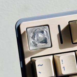 Könnte beinhalten: Nahaufnahme einer mechanischen Tastatur mit cremefarbenen Tastenkappen und einem hellblauen Rahmen. Die transparente, strukturierte Tastenkappe ist der Blickfang. Andere Tastenkappen zeigen Symbole und die Zahl 1.