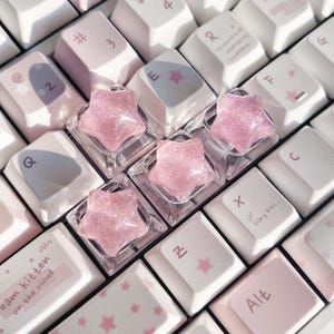 Könnte beinhalten: Nahaufnahme einer Tastatur mit weißen und rosa Tastenkappen. Vier transparente, sternförmige Tastenkappen mit rosa Glitzer liegen auf der Tastatur. Einige Tastenkappen haben rosa Sterne oder Text, darunter "dream kitten on the cloud."
