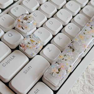 Puede incluir: Teclas de teclado de resina transparente con diseños florales y detalles de letras doradas. Las teclas están decoradas con pequeñas flores de colores en tonos rosa, amarillo y azul. Las teclas del teclado son blancas.