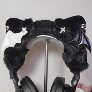 Op de afbeelding: Zwarte koptelefoon van imitatiebont met vleermuisvleugels en kruizen. De koptelefoon heeft een zachte textuur en wordt op een zilveren standaard gepresenteerd. Eén vleugel is wit, de andere is zwart.