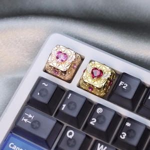 Pode incluir: Close-up de um teclado branco com teclas personalizadas. Duas teclas douradas são visíveis, cada uma adornada com uma pedra preciosa em forma de coração e detalhes intrincados. As teclas do teclado são pretas com letras brancas.