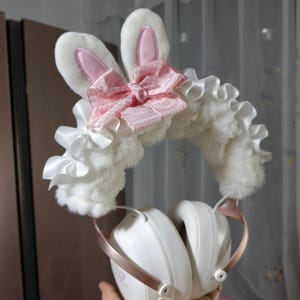 May include: White plush headphone cover featuring upright rabbit ears with pink centres, a pink and white gingham bow, and white satin ruffles. The cover is attached to white over-ear headphones with metallic rose gold accents on the frame.