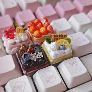 May include: Four miniature square cake-shaped keycaps featuring detailed toppings including strawberries, blueberries, lemon slices, kiwi, cream swirls, and small flowers, arranged on a mechanical keyboard with light pink and white keys.