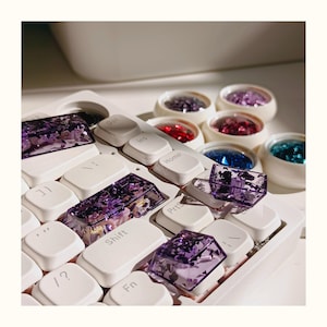 Puede incluir: Un teclado mecánico blanco con teclas personalizadas. Las teclas son de color púrpura transparente con purpurina y elementos florales incrustados. Pequeños recipientes redondos contienen purpurina de colores en varios tonos, incluyendo rojo, azul y púrpura.