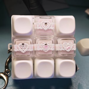 May include: A square grid of nine white keycaps arranged in three rows. The middle row features three transparent keycaps filled with light pink liquid, each wrapped in a white lace ribbon with a central heart-shaped charm and a small rhinestone.