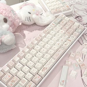 May include: A white mechanical keyboard featuring custom keycaps with pastel pink and light blue illustrations of cartoon characters and teddy bears. The keyboard includes a matching numeric keypad and several loose keycaps scattered nearby.