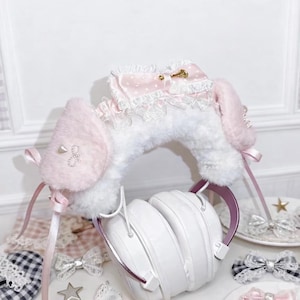 Può includere: Copricuffie in peluche bianco e rosa chiaro con una morbida texture in pelliccia sintetica, decorato con un fiocco in raso rosa a pois, un delicato bordo in pizzo bianco e piccoli accenti simili a perle sui lati, con lunghi nastri in raso.