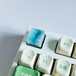 May include: A custom keyboard with a unique blue resin keycap. The keycap has a swirling blue design encased in clear resin. Other keycaps are white with green and black symbols. The keyboard is on a white surface.