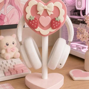 Puede incluir: Un soporte para auriculares blanco con una parte superior en forma de corazón decorada con un lazo color crema, dos fresas rojas y un corazón rosa más pequeño. El soporte tiene un poste vertical y una base sólida en forma de corazón rosa.