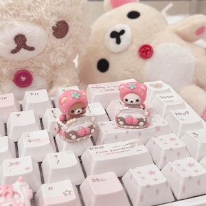 May include: Two small, sculpted bear-shaped keycaps featuring pink strawberry-themed hoods, each sitting in a miniature vehicle, placed on a white mechanical keyboard with pink star patterns and the text Love boundless on the spacebar key.