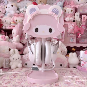 Pode incluir: Um suporte de fone de ouvido rosa com um design de personagem de desenho animado e fones de ouvido brancos. O suporte tem uma base redonda e um topo em forma de personagem com orelhas e laços. Os fones de ouvido são brancos com detalhes prateados.