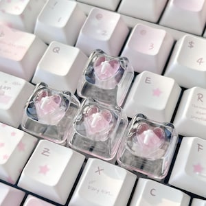 Puede incluir: Primer plano de un teclado con teclas personalizadas. Las teclas son transparentes con un diseño en forma de corazón rosa. Las teclas del teclado son blancas y rosas, algunas con estrellas rosas y texto.