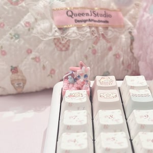 Puede incluir: Una tecla de teclado mecánico blanca con un pequeño personaje esculpido de color rosa y blanco, similar a un conejo, sentado en una cesta llena de objetos en miniatura. La superficie de la tecla muestra una ilustración impresa de un postre.
