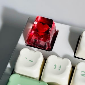 Puede incluir: Una tecla roja en forma de corazón se encuentra entre un conjunto de teclas blancas y verdes. La tecla del corazón es translúcida, con un corazón rojo en su interior. Las otras teclas tienen un diseño de oso con detalles verdes y números.