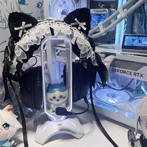 May include: A black cat ear headset accessory featuring white lace trim, small white bows with pearl accents, and long black ribbons. The headset is mounted on a white vertical stand with a small blue and white character figure inside the frame.