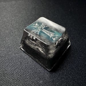 May include: A silver-colored, square-shaped keycap with a cross design on the top. The cross is detailed with small, sparkling accents, set against a teal background. The keycap has a textured, metallic appearance.