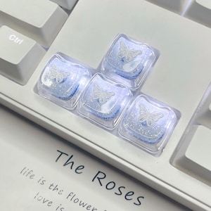 Pode incluir: Um conjunto de cinco teclas de teclado personalizadas. Cada tecla é quadrada e transparente, com um interior azul claro e brilhante. Um desenho de borboleta branca está centrado em cada tecla. O texto "The Roses" é visível.