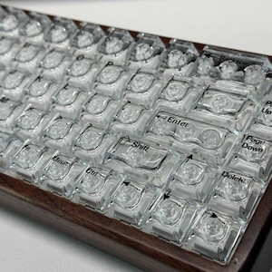 May include: A mechanical keyboard featuring a dark brown wooden frame and transparent, clear keycaps. The keys display black lettering for functions including Enter, Shift, Ctrl, Backspace, Delete, Page Up, and Page Down, revealing the internal switch mechanisms beneath each individual keycap.