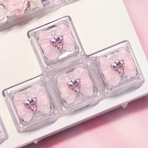 May include: Close-up of clear keyboard keycaps, each adorned with a light pink bow and a heart-shaped gem. The gems and bows are centered within the square keycaps, creating a delicate and feminine aesthetic. The background is a soft pink.