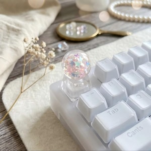 Peut inclure: Un capuchon de touche sphérique transparent rempli de confettis en forme d'étoiles iridescentes est placé sur un clavier blanc. Le capuchon de touche est un élément décoratif, ajoutant une touche de fantaisie au design du clavier. Le clavier a des touches blanches.