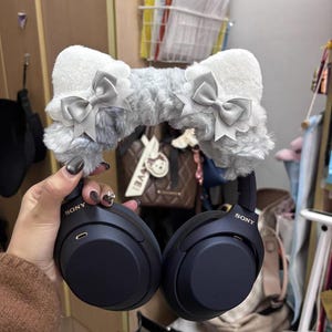 May include: Black Sony over-ear headphones featuring a plush, grey, faux-fur headband cover. The headband is adorned with two white, rounded cat-ear shapes, each decorated with a small, light grey satin ribbon bow on the front side.