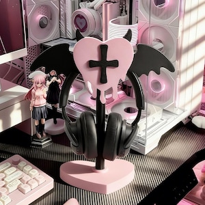 May include: A pink heart-shaped headphone stand with a black cross and dripping design, black horns, and bat wings. Black headphones rest on the stand. The stand is on a desk with a pink keyboard and computer components in the background.