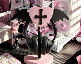 May include: A pink heart-shaped headphone stand with a black cross and dripping design, black horns, and bat wings. Black headphones rest on the stand. The stand is on a desk with a pink keyboard and computer components in the background.